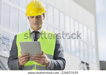 Young male engineer in reflector-vest and hardhat using digital tablet outside industry