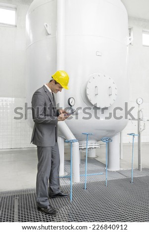 Full length side view of young male supervisor with clipboard standing by storage tank in industry