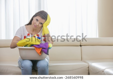 Portrait of tired woman with basket of cleaning supplies sitting on sofa at home