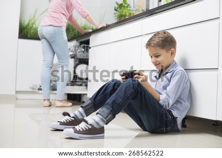 Full length of boy using hand-held video game with mother in background at kitchen