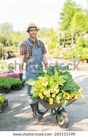 Full-length portrait of happy gardener pushing wheelbarrow with plants at garden