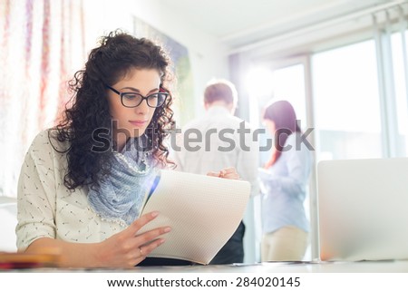 Businesswoman reading notepad at creative office with colleagues in background