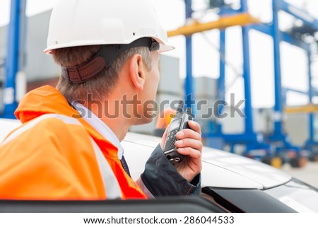 Middle-aged man using walkie-talkie while standing beside car in shipping yard