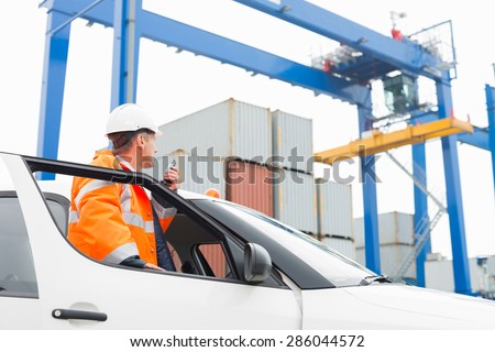 Middle-aged man using walkie-talkie while standing at car door in shipping yard