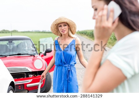 Angry woman looking at female using cell phone by damaged cars on road