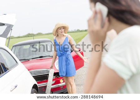 Angry woman standing by damaged cars with female using cell phone in foreground