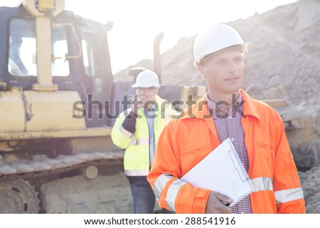 Engineer holding clipboard on construction site with colleague in background