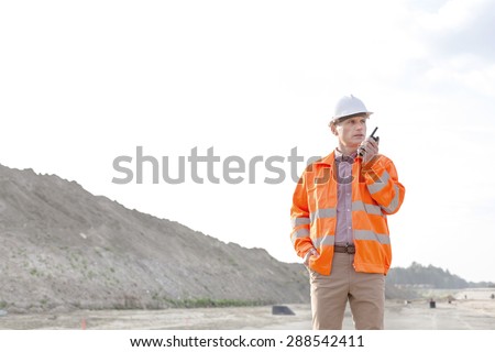Confident male supervisor using walkie-talkie on construction site against clear sky