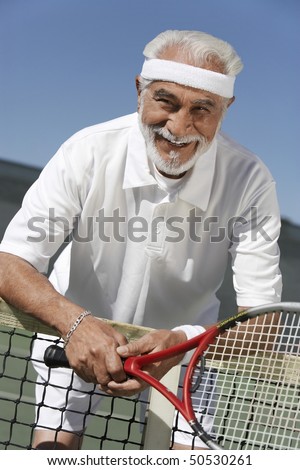 Smiling senior Man leaning on tennis net, holding tennis racket