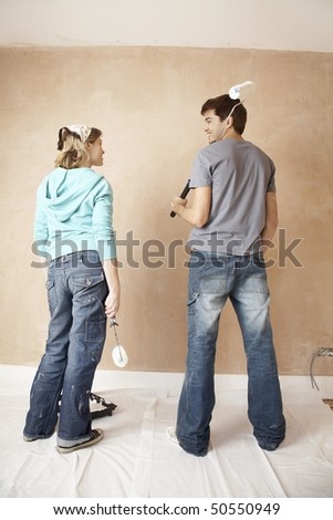 Couple standing, holding paint rollers in unrenovated room, back view