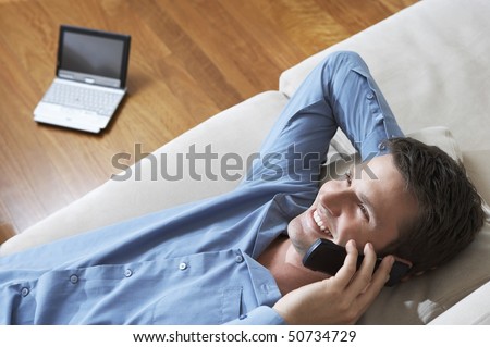 Man lying on sofa using mobile phone, laptop in background, view from above.