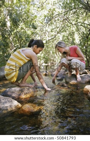 Two teenage girls (16-17 years) squatting on stone by stream in forest, hands in water
