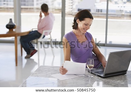 Woman using laptop sitting at table, in background man sitting at table on mobile phone