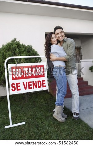 Couple standing in front of house with For Sale sign, portrait