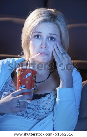 Young woman sitting in theatre, drinking soft drink, hand on cheek, watching sad movie