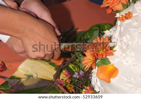 Two hands holding a knife cutting a wedding cake with flowers on top.