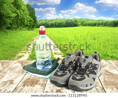 Sport shoes, towel and water in bottle on wooden boards and field with sky on background