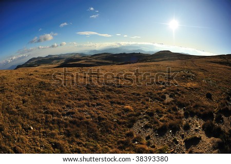Fish eye shot of mountain landscape with brown grass and setting sun with hills in background and white clouds