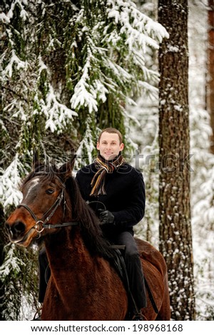 Young attractive man riding black horse at white winter forest.