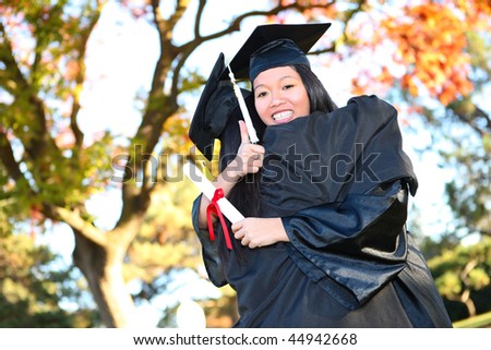 Pretty Asian woman wearing cap and gown holding diploma at graduation hugging friend