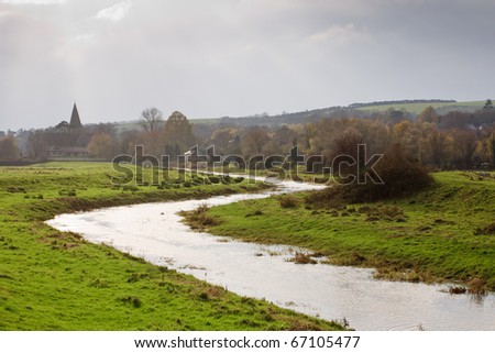 A meandering river through the green meadows to the country village in the background