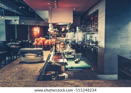 Restaurant kitchen interior: bar counter made of natural stone, fences off the open kitchen and hall for visitors with tables and chairs. In the background buzzing restaurant work motion chefs