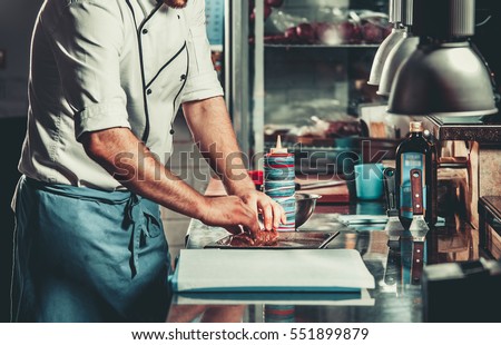 Young white chef in kitchen interior. Man marinating beef steak on a tray. Meat ready for the grill and serve. Only hands