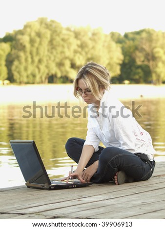 A young girl using a laptop on a wooden river embankment.