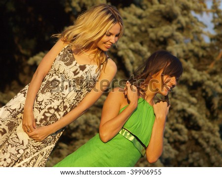 A young blond and a brunette wearing sundresses in front of fur-trees.