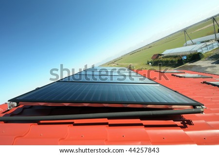 Close up of red roof with solar panel. Blue sky, green field and greenhouse in background.