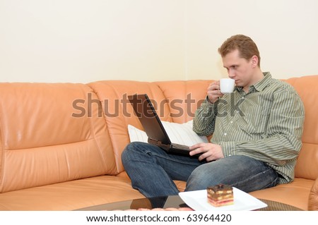 Young man drinking coffee and working on portable computer on leather sofa in living room.