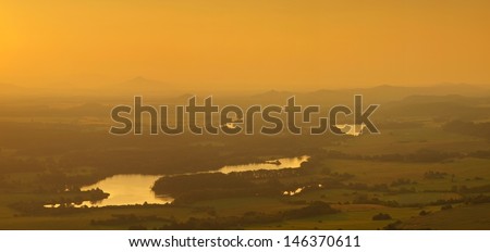 View into foggy countryside with bushes, forests, villages, levels of ponds within early morning sunrise.