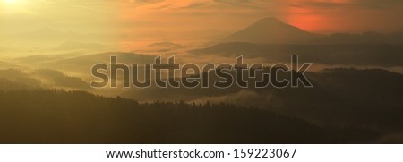 Sunrise in a beautiful mountain of Bohemian-Saxony Switzerland. Sandstone peaks and hills increased from foggy background, the fog is orange due to sun rays.