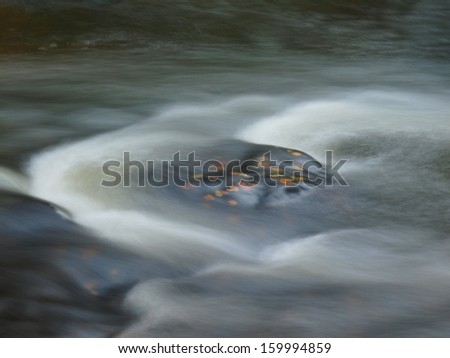 Orange beech leaves on mossy stone below increased water level. Blurred motion of waves around the stone.
