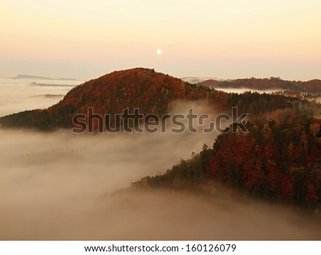 Full moon night with sunrise in a beautiful mountain of Bohemian-Saxony Switzerland. Sandstone peaks and hills increased from foggy background, the fog is orange due to sun rays.