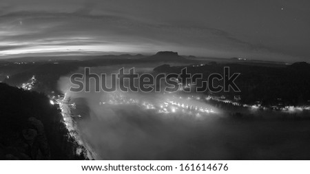 Early morning at river after rainy night, fog cloud is shaking above water level and river banks. Autumn landscape, daybreak at horizon. Traffic lights are shining in the mist. Black and white photo.