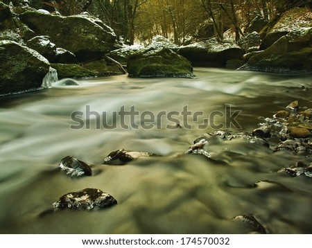 Dark green cold water of mountain stream in winter time between big boulders with snowflakes of first powder snow.