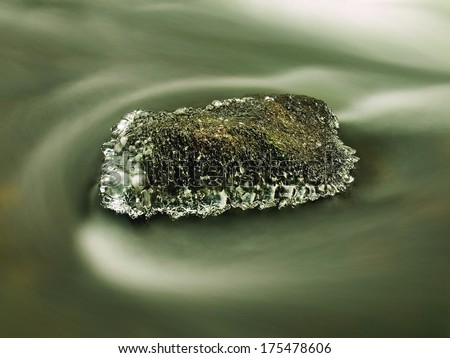 Freeze mossy sandstone boulder in dark cold water of mountain river. Small icicles glitter above milky blurred water of stream.