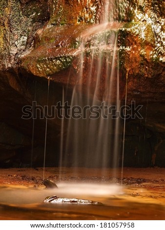 Small waterfall on small mountain stream, mossy sandstone block and water is jumping down into small pool. Water streams with sun rays, orange sand with sediments below.