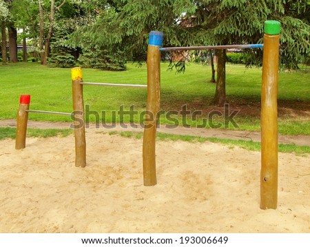 Steel horizontal bars on wooden pillars in children playground. Orange sand below bars, green park in the background.