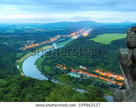 Early spring morning above big river after rainy night, fresh blue cloud in the sky, lights in town on river banks. Spring green forests in landscape, daybreak at horizon