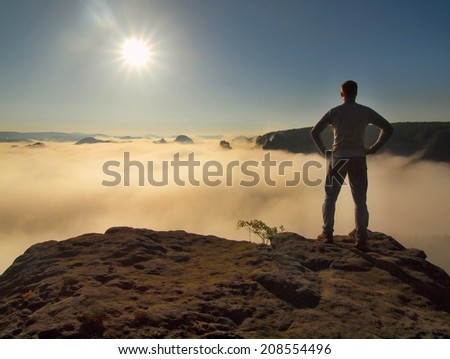 Man in shirt and jeans is standing on the peak of sandstone rock in national park Saxony Switzerland and watching over the misty and foggy morning valley to Sun. Beautiful moment the miracle of nature