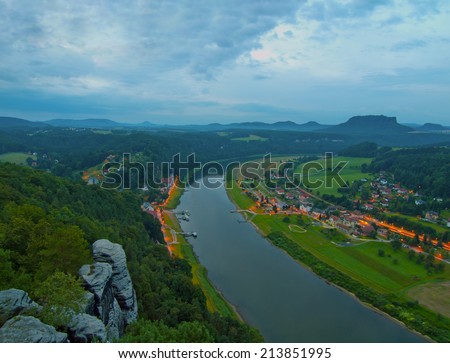 Early autumn morning on rocks above big river, fresh blue cloud in the sky, lights in town on river banks. Green forests in landscape, daybreak at horizon