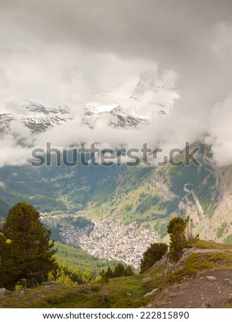 Deep valley below misty peaks of Alps mountains in background. Small mountain town under mist cover