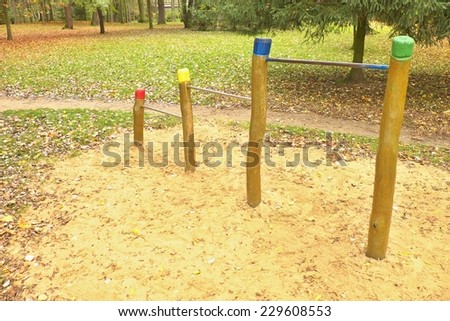 Steel horizontal bars on wooden pillars in children playground. Orange sand below bars, green park in the background.