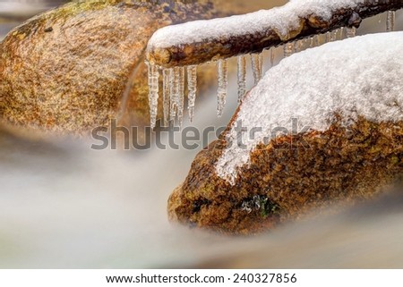 Smoky cold water of mountain stream in winter time. Long crystal icicles are hanging above milky water level. Big granite boulders.