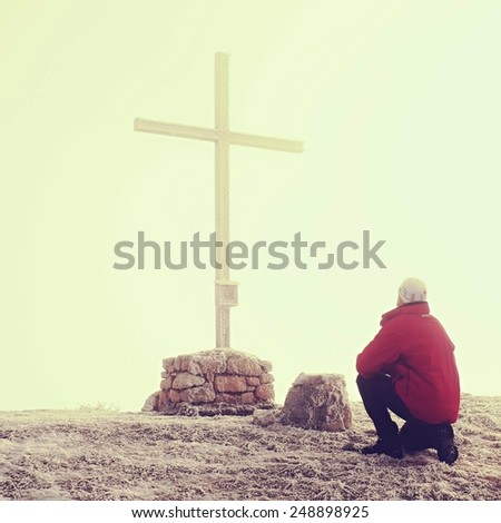 Tourist in red is kneeling at cross memorial on mountain peak. Misty Alpine winter day. Metal cross at a mountain peak.