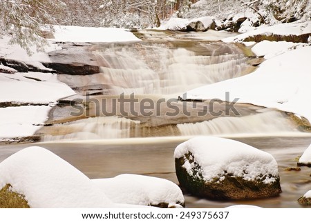 Winter at mountain river. Big stones in stream covered with fresh powder snow and lazy water with low level. Reflections of forest in water level.