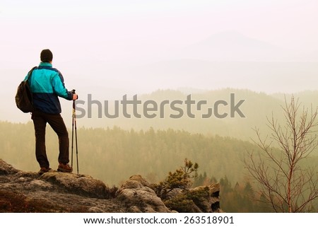 Tourist in blue green windcheater with sporty backpack and poles in hands stand on rocky view point and watching into deep misty valley bellow. Misty spring daybreak in rocky mountains.