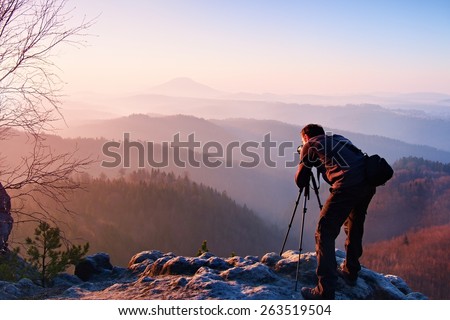 Professional photographer takes photos with camera on tripod on rocky peak. Dreamy fogy landscape, spring orange pink misty sunrise in a beautiful valley below
